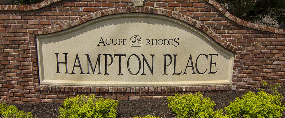 The brick entrance sign for Hampton Place, set in a curved wall above a bed of green shrubs.