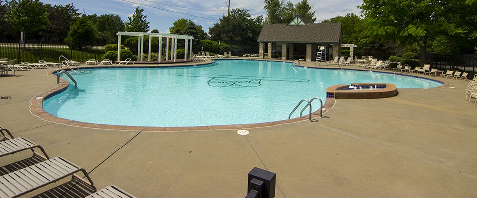 The community pool on a sunny day, with lounge chairs around the deck, a white pergola and the pool house behind it.
